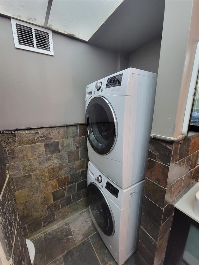 laundry area featuring tile walls, stone tile floors, laundry area, and stacked washer / dryer