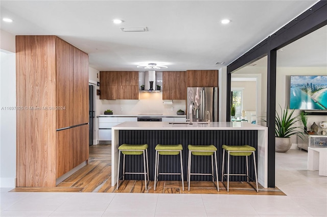 kitchen with wall chimney exhaust hood, stainless steel fridge, light hardwood / wood-style floors, a kitchen bar, and a center island with sink