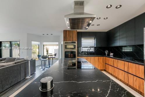 kitchen with island exhaust hood, decorative backsplash, black electric cooktop, double oven, and dark stone countertops