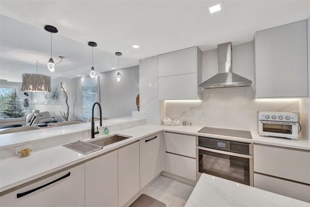 kitchen featuring pendant lighting, wall chimney range hood, sink, black electric cooktop, and oven