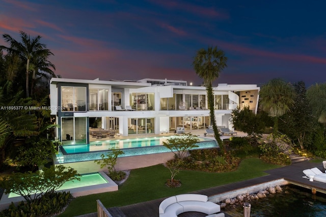 back house at dusk featuring a swimming pool with hot tub, a patio, a balcony, and a yard