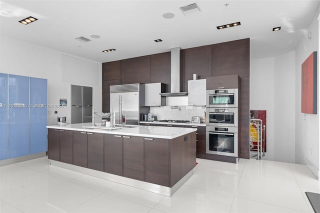 kitchen featuring a center island with sink, wall chimney exhaust hood, stainless steel appliances, and light tile patterned floors