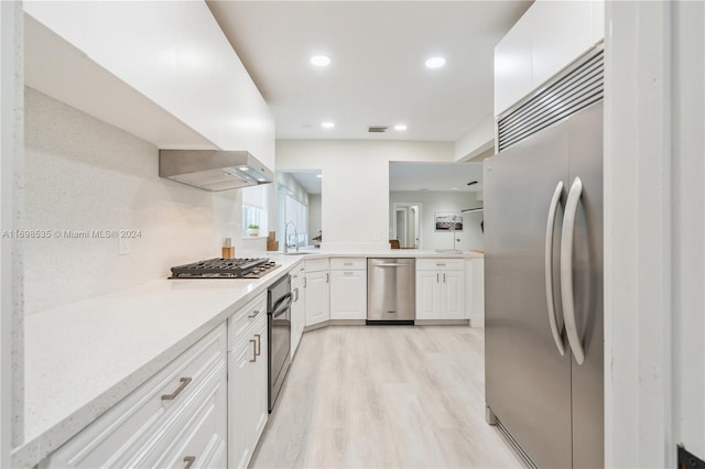 kitchen featuring appliances with stainless steel finishes, light stone counters, wall chimney exhaust hood, white cabinets, and light hardwood / wood-style floors