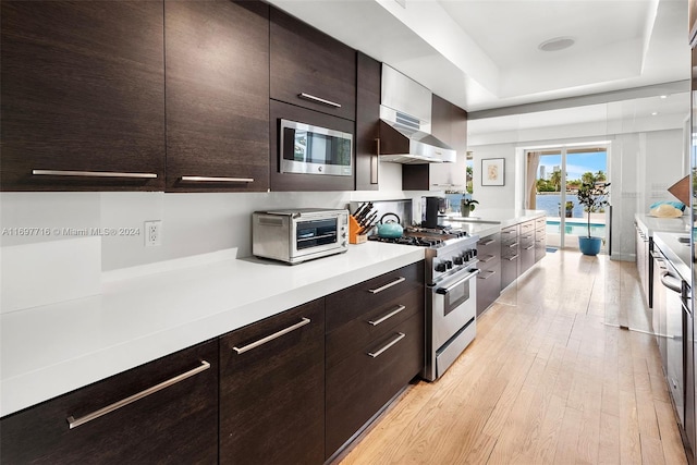 kitchen featuring wall chimney exhaust hood, a tray ceiling, dark brown cabinets, light hardwood / wood-style floors, and stainless steel appliances