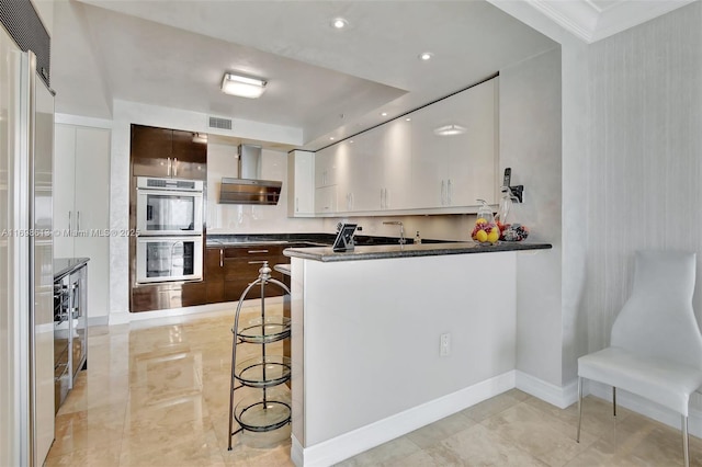 kitchen with wall chimney exhaust hood, kitchen peninsula, dark stone counters, a breakfast bar, and appliances with stainless steel finishes