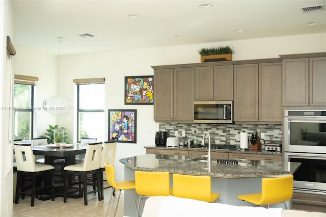 kitchen with tasteful backsplash, dark stone countertops, a kitchen island with sink, light tile patterned flooring, and appliances with stainless steel finishes