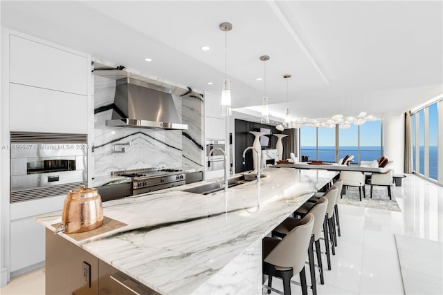 kitchen with hanging light fixtures, wall chimney range hood, a water view, decorative backsplash, and white cabinets