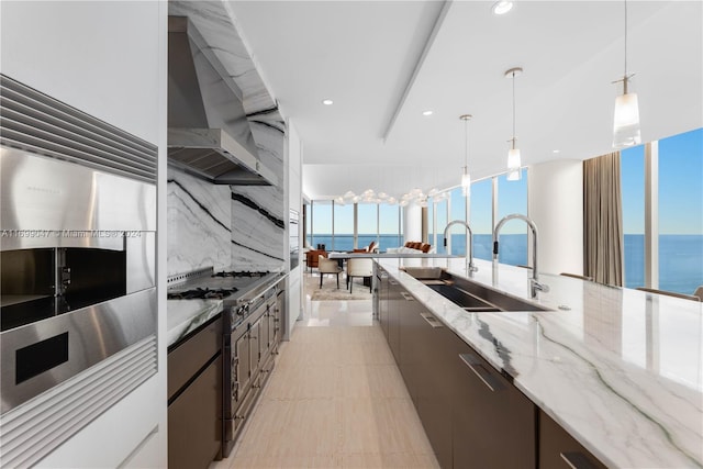 kitchen with sink, wall chimney exhaust hood, hanging light fixtures, light stone counters, and a water view