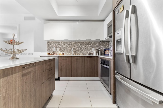 kitchen with white cabinetry, a tray ceiling, decorative backsplash, light tile patterned flooring, and appliances with stainless steel finishes