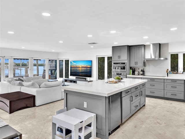 kitchen with wall chimney exhaust hood, gray cabinetry, double oven, stovetop, and a center island