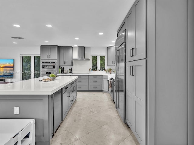 kitchen with gray cabinetry, wall chimney exhaust hood, stainless steel appliances, and a kitchen island