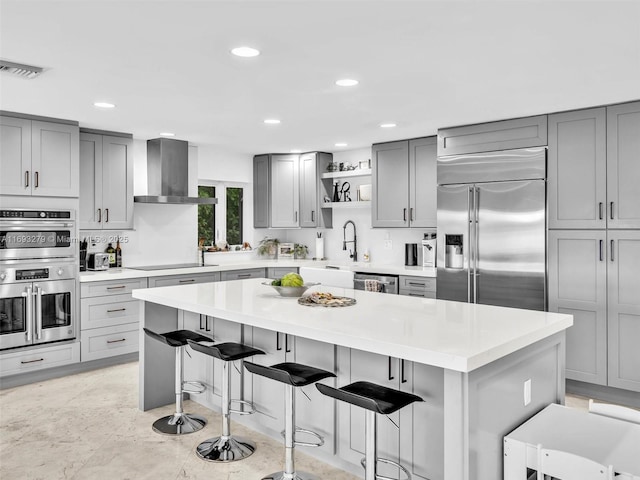 kitchen featuring a center island, wall chimney range hood, a kitchen breakfast bar, gray cabinets, and appliances with stainless steel finishes