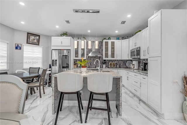 kitchen featuring white cabinets, wall chimney exhaust hood, light stone countertops, an island with sink, and stainless steel appliances