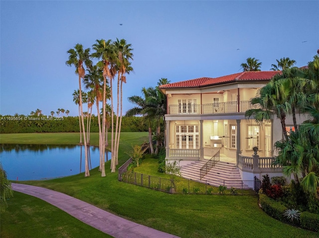 back house at dusk with a lawn, a balcony, and a water view