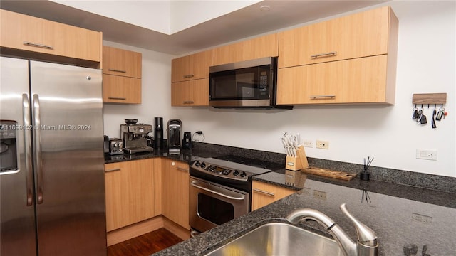 kitchen with appliances with stainless steel finishes, light brown cabinetry, sink, hardwood / wood-style flooring, and dark stone countertops