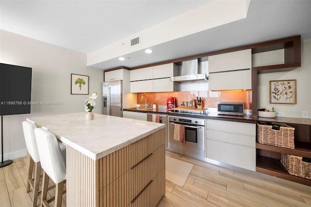 kitchen featuring wall chimney exhaust hood, a kitchen island, appliances with stainless steel finishes, white cabinets, and light wood-type flooring