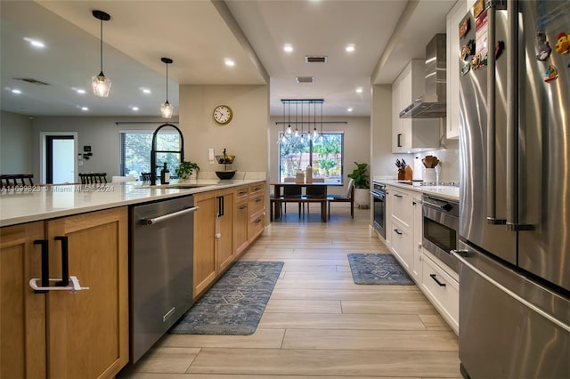 kitchen with plenty of natural light, white cabinets, wall chimney range hood, appliances with stainless steel finishes, and decorative light fixtures