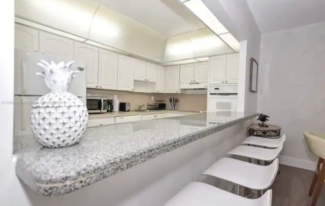 kitchen with dark wood-type flooring, white oven, kitchen peninsula, light stone countertops, and white cabinetry