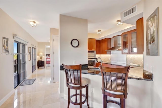 kitchen featuring decorative backsplash, a kitchen bar, double oven, sink, and wall chimney range hood