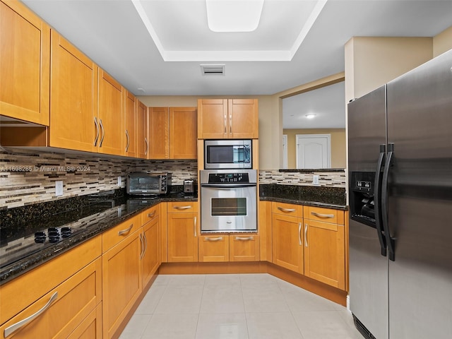 kitchen featuring dark stone countertops, appliances with stainless steel finishes, tasteful backsplash, a tray ceiling, and light tile patterned flooring