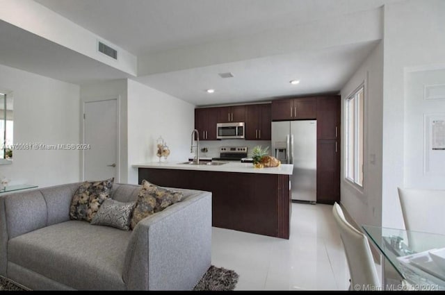 kitchen with dark brown cabinetry, stainless steel appliances, a kitchen island with sink, sink, and light tile patterned floors