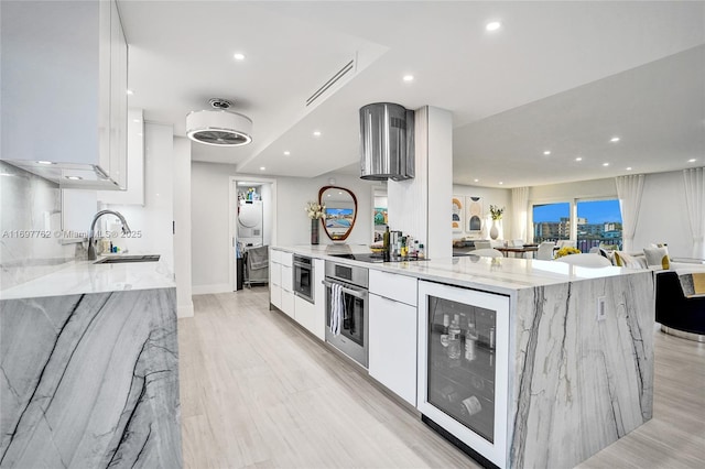 kitchen with wine cooler, stainless steel oven, light stone countertops, white cabinets, and sink