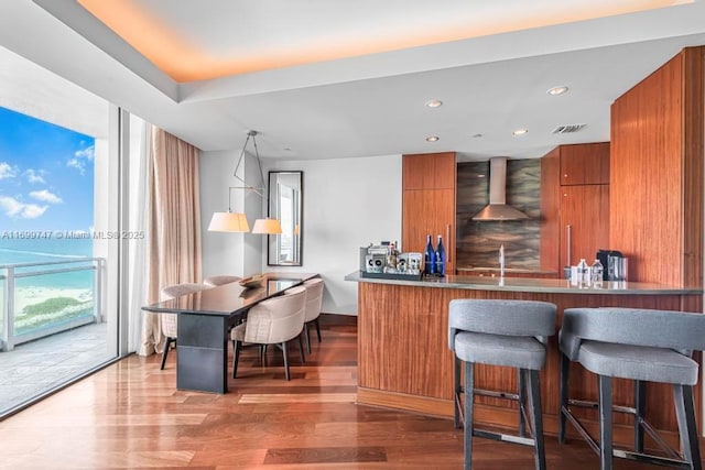 kitchen featuring wood-type flooring, wall chimney range hood, kitchen peninsula, and a breakfast bar area