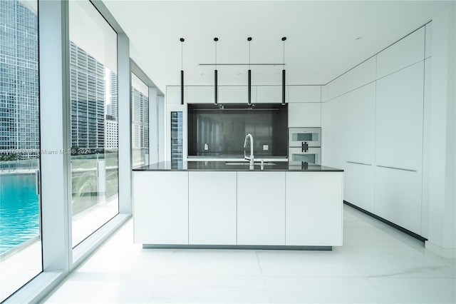 kitchen featuring pendant lighting, backsplash, white cabinets, a center island with sink, and sink