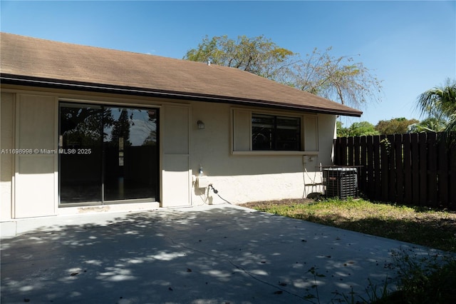 exterior space featuring fence, roof with shingles, stucco siding, cooling unit, and a patio