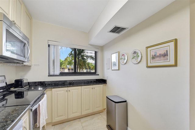 kitchen featuring stainless steel appliances, light tile patterned flooring, cream cabinetry, and dark stone countertops