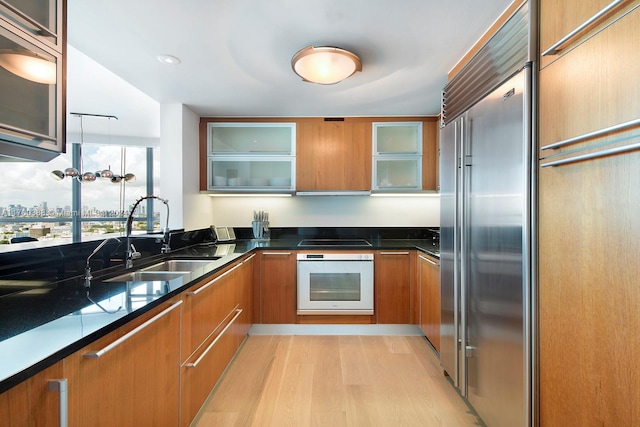 kitchen with white oven, sink, light hardwood / wood-style flooring, dark stone countertops, and stainless steel built in refrigerator