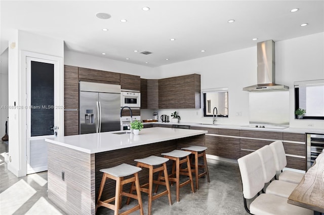 kitchen with sink, wall chimney range hood, built in refrigerator, a kitchen island with sink, and a breakfast bar