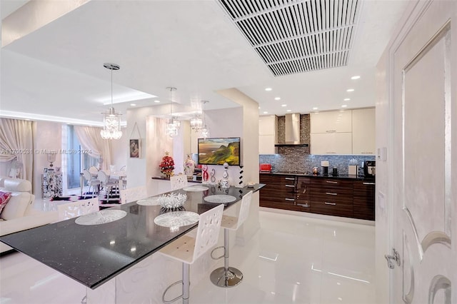 kitchen featuring wall chimney range hood, tasteful backsplash, a kitchen bar, dark brown cabinets, and light tile patterned floors