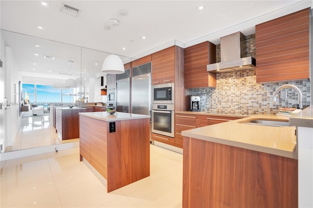 kitchen featuring wall chimney range hood, sink, hanging light fixtures, built in appliances, and a kitchen island