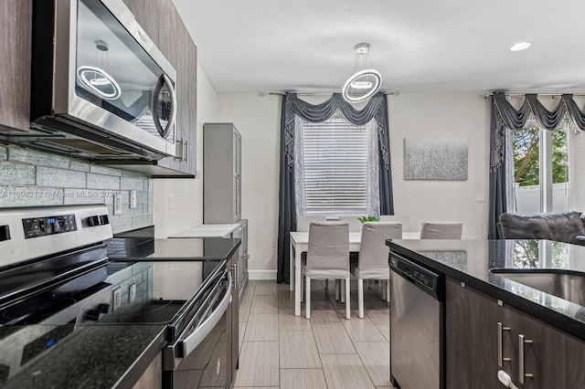 kitchen featuring decorative backsplash, appliances with stainless steel finishes, dark brown cabinets, dark stone countertops, and hanging light fixtures