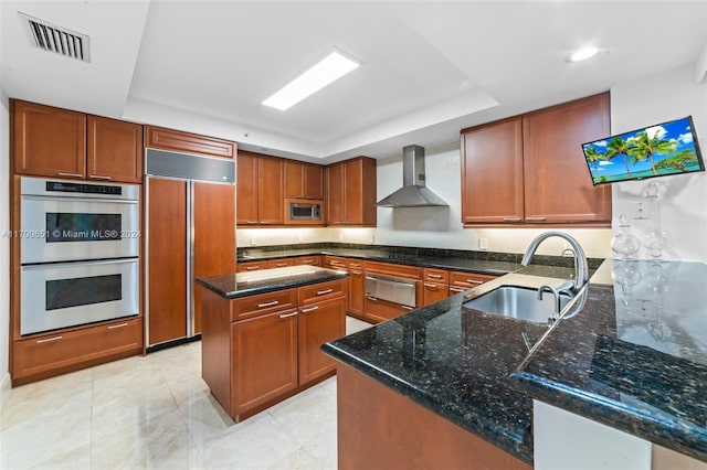 kitchen featuring sink, wall chimney exhaust hood, built in appliances, dark stone countertops, and a tray ceiling