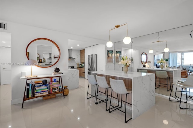 kitchen with a center island, hanging light fixtures, wall chimney exhaust hood, stainless steel fridge, and light stone countertops