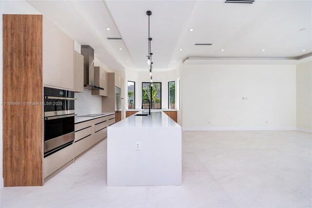 kitchen with sink, wall chimney exhaust hood, backsplash, decorative light fixtures, and a center island with sink