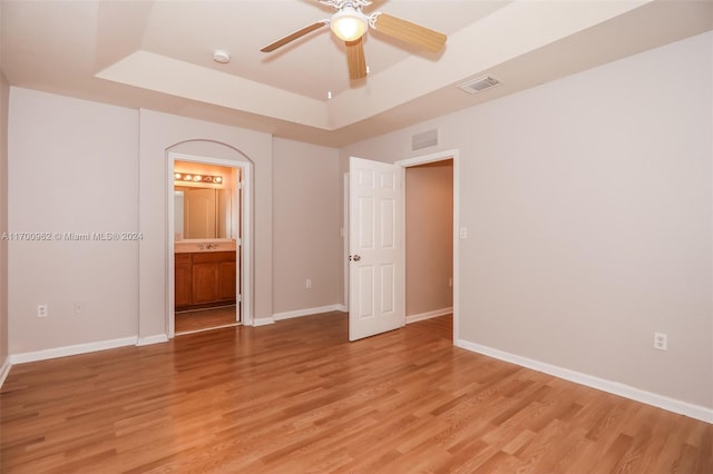 unfurnished bedroom with a raised ceiling, ensuite bathroom, ceiling fan, and light hardwood / wood-style floors