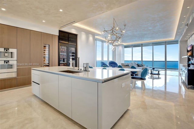 kitchen with a center island with sink, expansive windows, sink, a notable chandelier, and white cabinetry