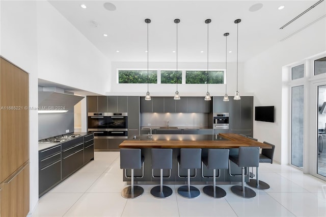 kitchen with wall chimney exhaust hood, sink, decorative light fixtures, a high ceiling, and an island with sink