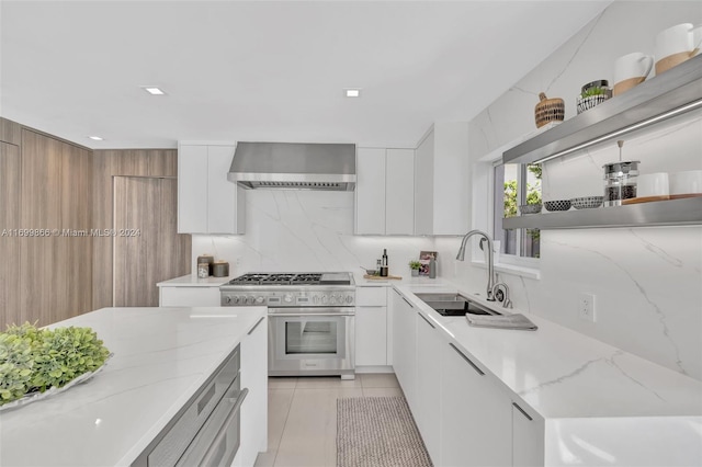 kitchen featuring wall chimney range hood, sink, light stone countertops, white cabinets, and range with two ovens