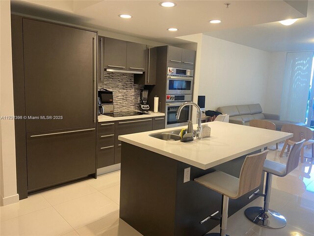 kitchen featuring a breakfast bar, a kitchen island with sink, backsplash, black electric cooktop, and double oven