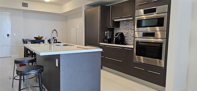 kitchen featuring double oven, a breakfast bar, a sink, backsplash, and a center island with sink