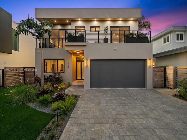 contemporary house featuring stucco siding, fence, decorative driveway, a balcony, and an attached garage