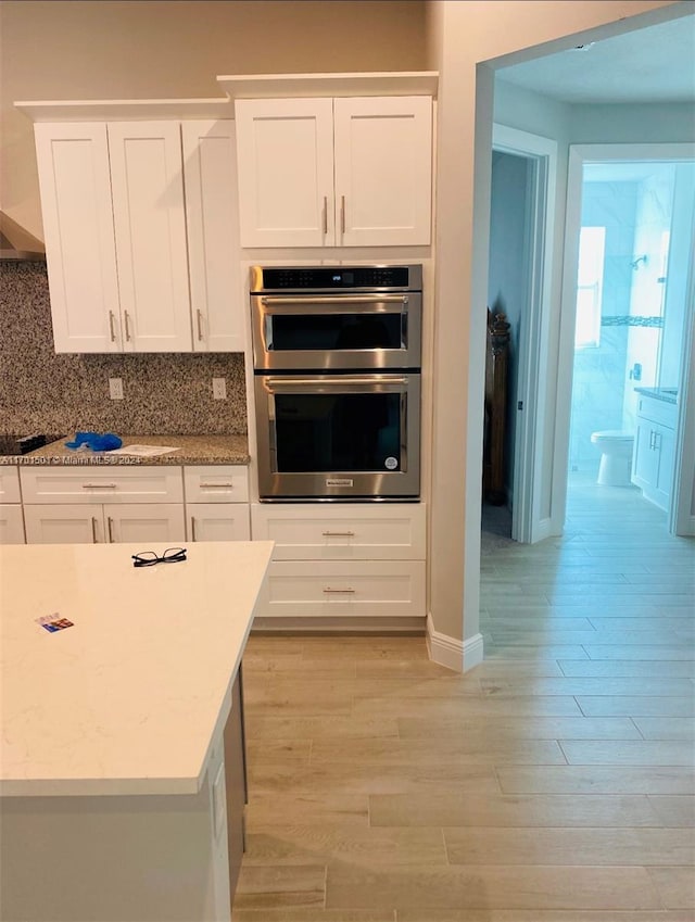 kitchen with double oven, tasteful backsplash, light hardwood / wood-style floors, light stone counters, and white cabinetry