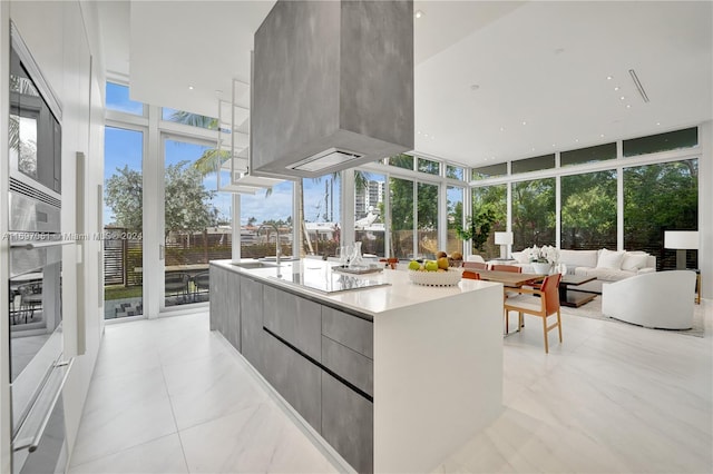 kitchen featuring floor to ceiling windows, gray cabinetry, stainless steel oven, a kitchen island with sink, and sink