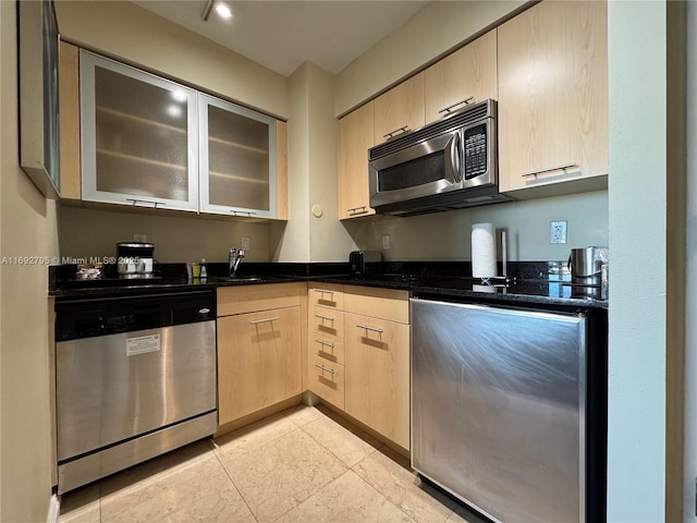 kitchen with stainless steel appliances, a sink, dark stone counters, light brown cabinetry, and glass insert cabinets