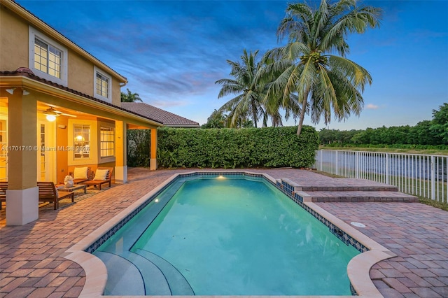 pool at dusk with a patio area, ceiling fan, and a water view