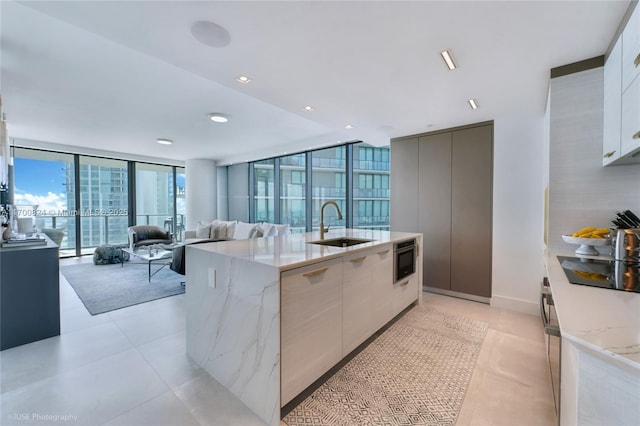 kitchen with light stone counters, expansive windows, black electric cooktop, sink, and an island with sink